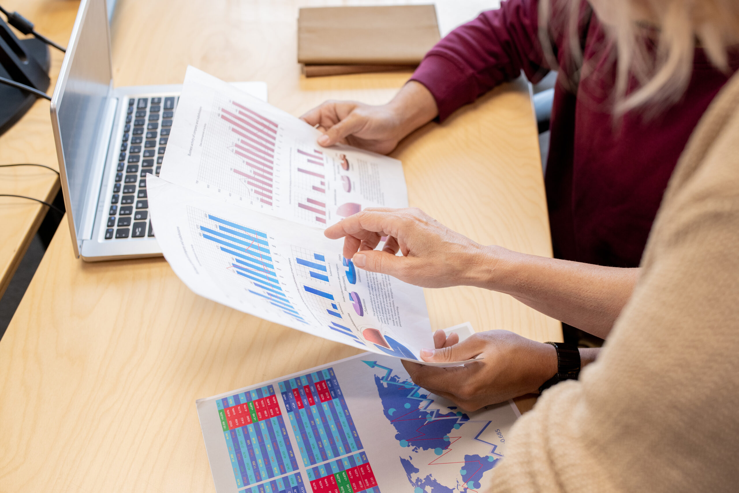 Hand of contemporary female broker pointing at one of financial papers held by colleague while discussing chart and graph by desk