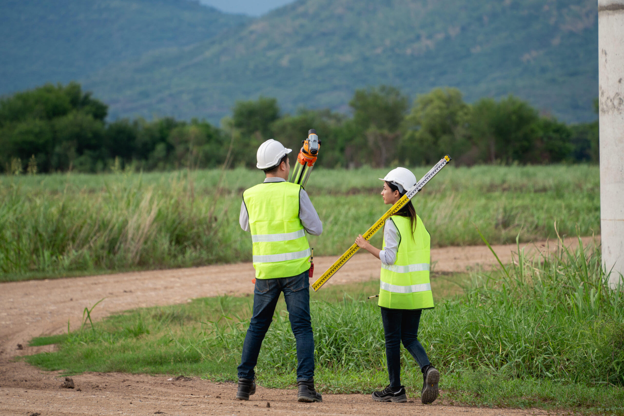 Two engineers wearing safety vests and holding surveying equipment. One of them is holding a yellow tape measure